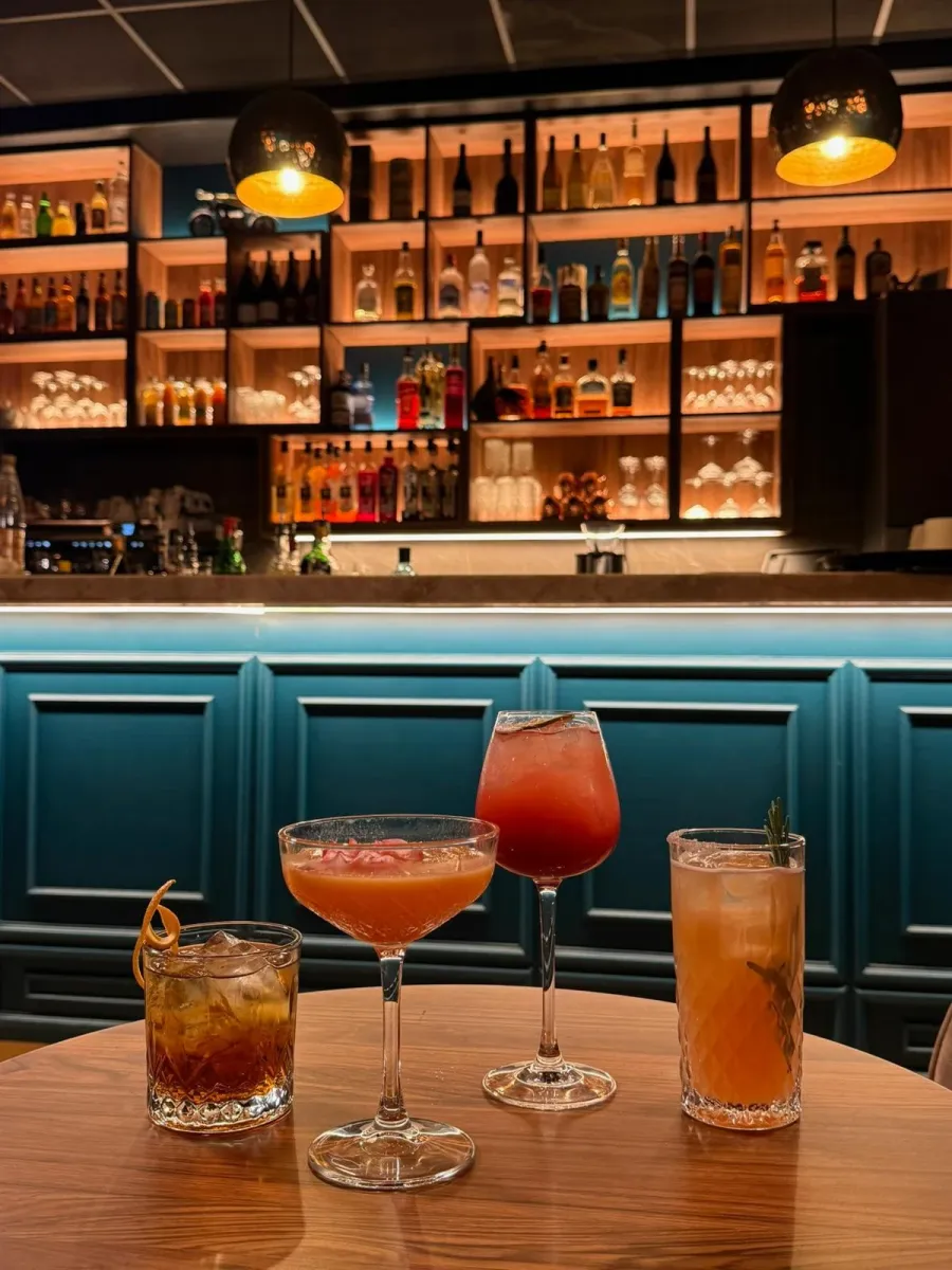 Quatre cocktails sur une table ronde, devant le comptoir et ses étagères de bouteilles